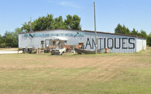 A large metal building with "ANTIQUE" painted on the side, surrounded by grass and a few old vehicles.