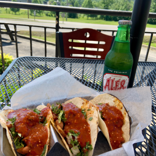 Three tacos topped with salsa and herbs, served with a green soda on a table outdoors.