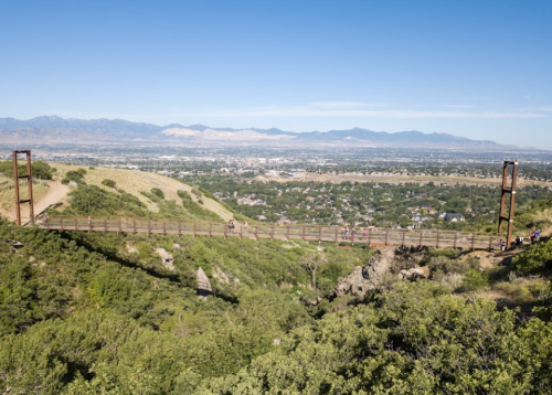 A suspension bridge spans a canyon, surrounded by greenery and mountains under a clear blue sky.