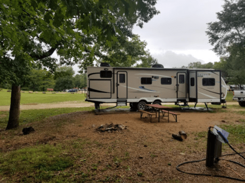 A parked RV near a picnic table and fire pit, surrounded by trees and grassy area on a cloudy day.