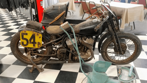 An old, rusty motorcycle with a yellow number plate, parked on a checkered floor in a room with tables and chairs.