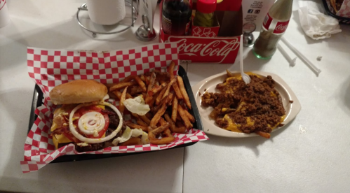 A tray with a burger, fries, and a plate of chili cheese fries on a table with condiments in the background.