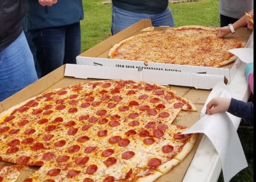 Two large pizzas on a table: one with pepperoni and one with cheese, surrounded by people.