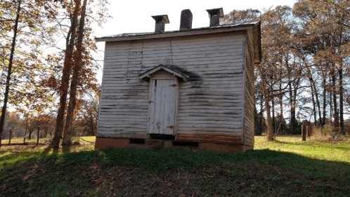 A weathered wooden building with a small door, surrounded by trees and grass in a rural setting.