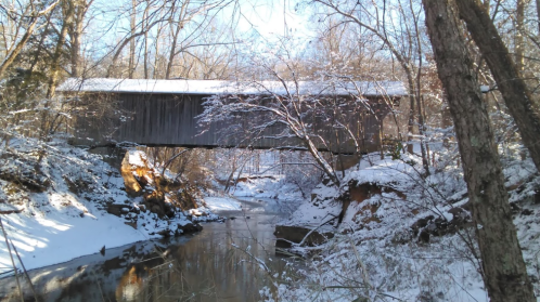 A snow-covered landscape featuring a wooden covered bridge over a calm stream, surrounded by trees.