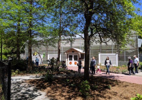 A group of people walks past a greenhouse surrounded by trees and garden paths on a sunny day.