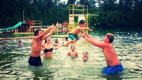 A group of children and teens play in a lake, with one boy jumping into the water while others cheer and catch him.
