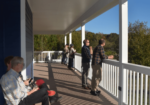 A group of people stands on a balcony, enjoying the view on a sunny day, with trees in the background.