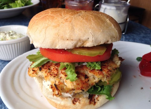 A close-up of a grilled chicken sandwich with lettuce, tomato, pickles, and mayonnaise on a toasted bun.