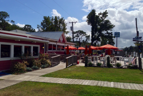 A colorful waterfront restaurant with outdoor seating and orange umbrellas, surrounded by greenery and a wooden walkway.