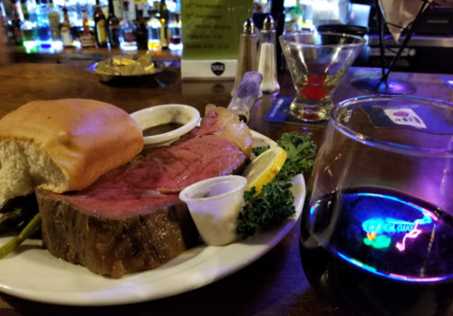 A plate of prime rib with sides, a roll, and a glass of red wine, set against a bar with bottles in the background.