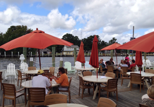 Outdoor dining area with orange umbrellas, wooden tables, and a view of the water, featuring people enjoying their meals.