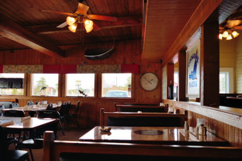 Cozy restaurant interior with wooden walls, ceiling fans, and tables set for dining, featuring large windows and a clock.