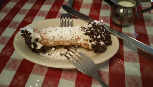 A cannoli filled with cream, topped with powdered sugar and chocolate chips, served on a plate with utensils.