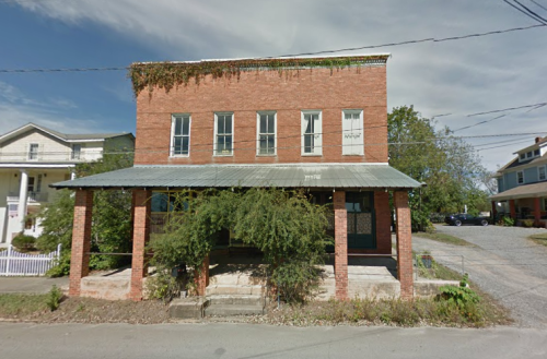 A two-story brick building with a porch, overgrown plants, and a metal roof, set in a rural area.
