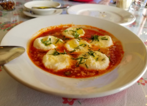A bowl of dumplings in a rich red sauce, garnished with fresh herbs, served on a floral tablecloth.
