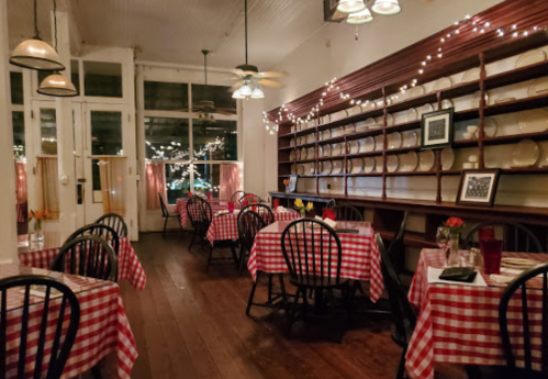 Cozy restaurant interior with checkered tablecloths, wooden floors, and warm lighting, featuring a shelf of plates.