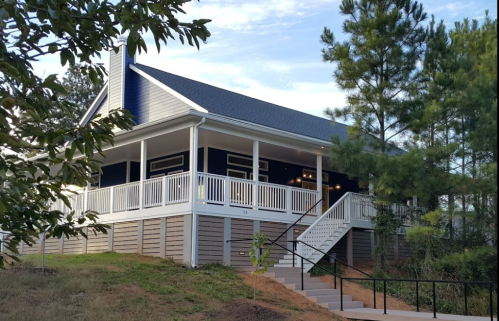 A modern house with a porch, surrounded by trees, set on a slight hill under a clear sky.