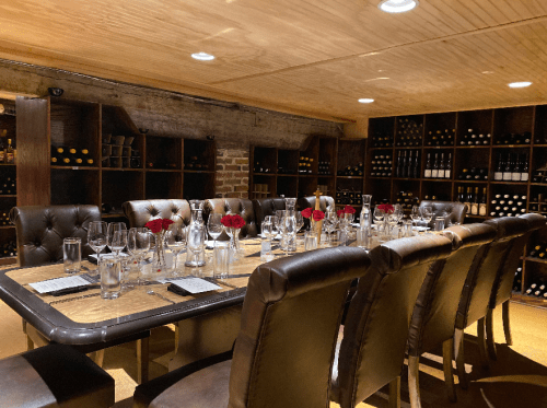 A dimly lit wine cellar with a long dining table set for a meal, surrounded by shelves of wine bottles.