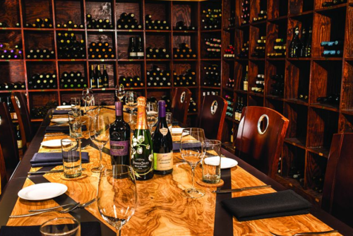 A dining table set with glassware and plates, surrounded by a wine cellar filled with bottles.