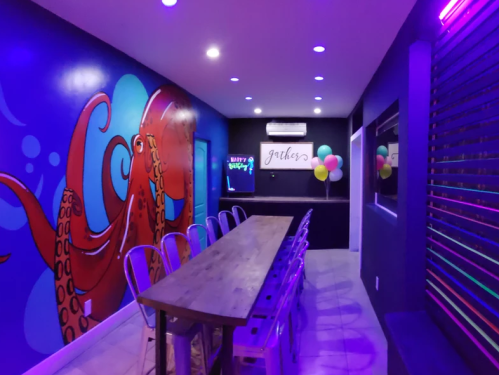 A vibrant room with purple lighting, featuring an octopus mural and a long wooden table with metal chairs.