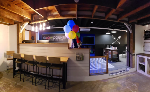 A brightly lit indoor mini-golf reception area with a counter, balloons, and a cozy seating arrangement.