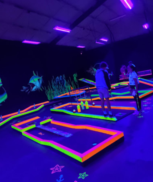 A vibrant indoor mini-golf course with glowing neon colors and blacklight effects, featuring players enjoying the game.