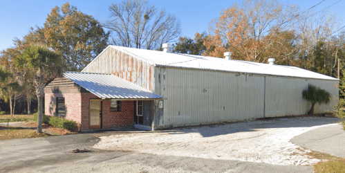 A large, industrial-style building with a metal roof and brick facade, surrounded by trees and a gravel driveway.
