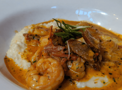 A creamy dish featuring shrimp, sausage, and vegetables served over grits, garnished with green herbs.