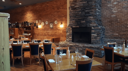 Cozy restaurant interior with wooden tables, blue chairs, and a stone fireplace against a brick wall.
