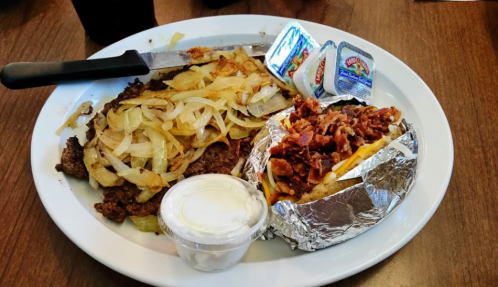 A plate with grilled meat topped with onions, a baked potato with bacon, and two cups of dipping sauce.
