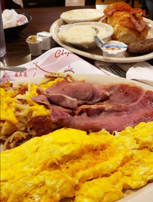 A hearty breakfast plate featuring ham, scrambled eggs, cheesy hash browns, and sides of biscuits and gravy.
