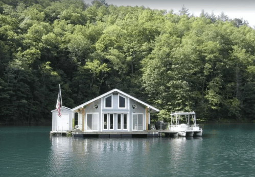 A modern houseboat on calm water, surrounded by lush green trees and featuring a small dock.