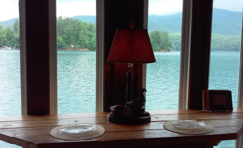 A cozy table with a lamp and plates, overlooking a serene lake and mountains through large windows.