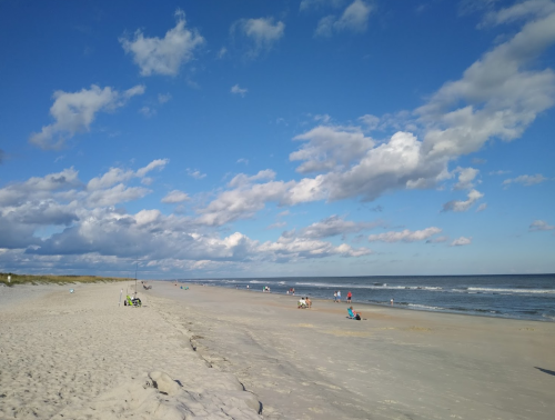 A sandy beach with people enjoying the sun, gentle waves, and a blue sky filled with fluffy clouds.