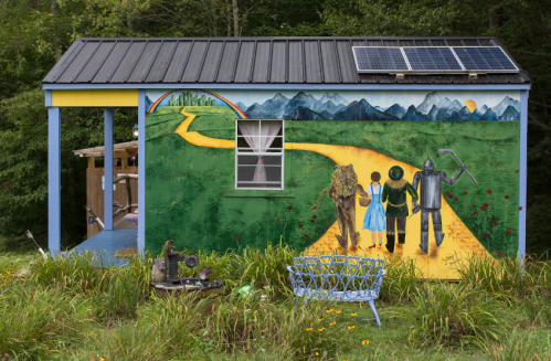 A colorful mural on a small building depicts characters walking a yellow brick road with mountains and a rainbow in the background.