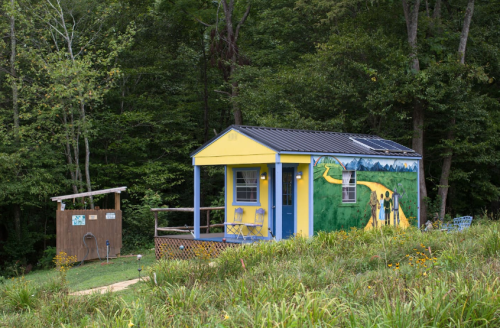 A colorful yellow cabin with a mural, surrounded by greenery and a small outdoor shower area nearby.