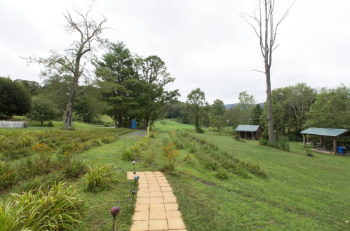 A scenic view of a grassy landscape with a stone path, trees, and two small structures in the distance under a cloudy sky.