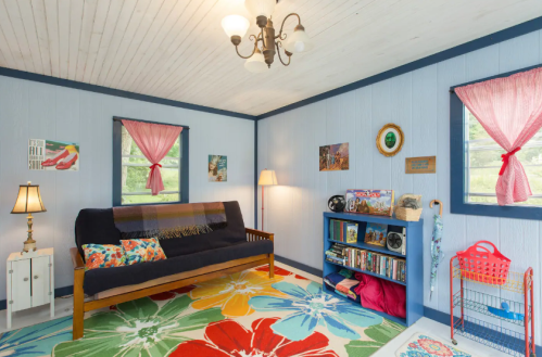 Cozy room with blue walls, colorful rug, a futon, lamp, bookshelf, and bright curtains. Decorated with art and books.