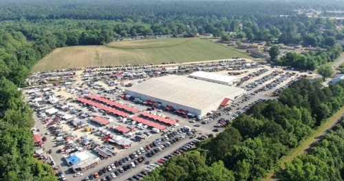 Aerial view of a large outdoor market with numerous cars and vendor tents surrounded by greenery.