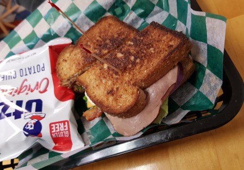 A toasted sandwich with layers of meat and lettuce, served with a bag of potato chips on a green checkered paper.