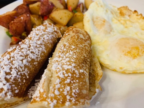 A plate with two powdered sugar-covered crepes, fried eggs, and seasoned diced potatoes.