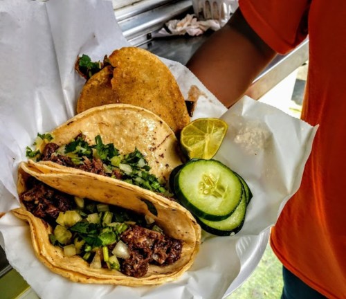 A plate of tacos with beef, cilantro, and onions, accompanied by cucumber slices and lime wedges.