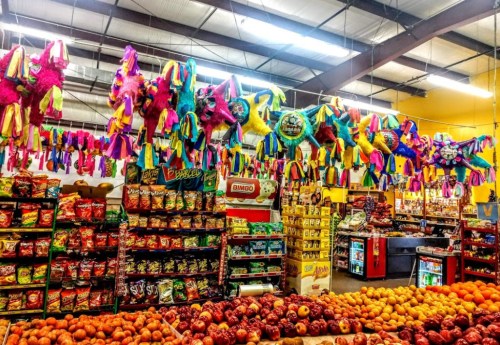 Colorful piñatas hang from the ceiling of a market, with shelves of snacks and fresh fruit below.