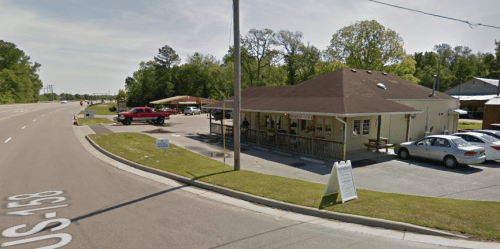 A small building with a porch and cars parked outside, located along a road with trees in the background.