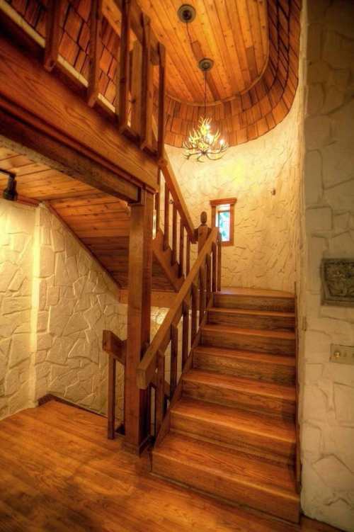 A wooden staircase with a rustic railing, leading to a second floor, surrounded by stone walls and warm lighting.