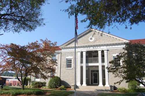 A stone building with a "City Hall" sign, surrounded by trees and a flagpole, under a clear blue sky.