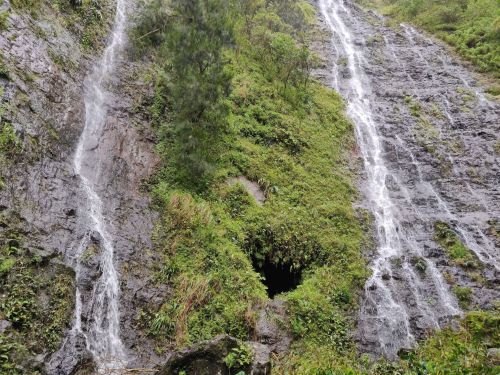Two waterfalls cascade down a rocky, green hillside, surrounded by lush vegetation and a dark cave entrance.