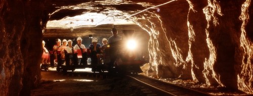 A small train with passengers enters a dimly lit tunnel, surrounded by rocky walls. Bright light shines from the front.