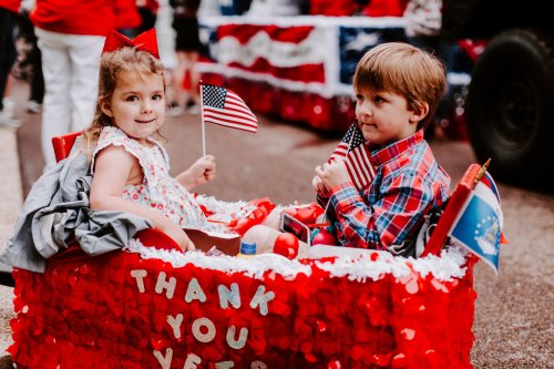Two children in a decorated wagon holding small American flags, smiling during a festive parade. Red, white, and blue decorations.